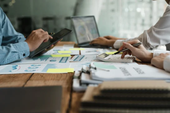 Profissionais tentando aprender como gerenciar os recursos financeiros da escola. Estão debruçados sobre uma mesa cheia de papéis, computador e calculadora.