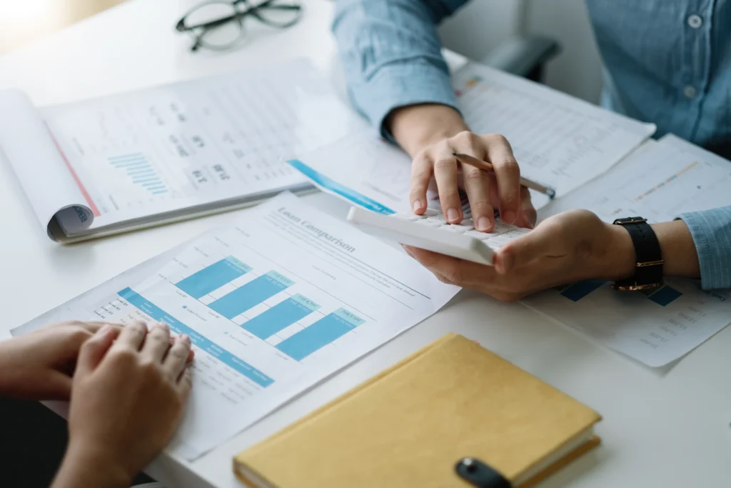 Papéis com gráficos e folhas em cima da mesa. Mãos seguram calculadora, enquanto uma pessoa explica o que é gestão de financeira escolar para outra.