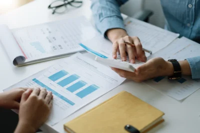 Papéis com gráficos e folhas em cima da mesa. Mãos seguram calculadora, enquanto uma pessoa explica o que é gestão de financeira escolar para outra.