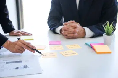 Homem debruçado sobre mesa com braços cruzados, no canto direito. À direita, mulher aponta com caneta para papéis em cima da mesa, mostrando como fazer gestão financeira em escritório de advocacia.