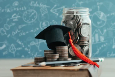 Chapéu de formatura e um pote de moedas em cima de mesa, com lousa escolar atrás. A foto representa a Lei da mensalidade escolar e o pagamento das mensalidades.