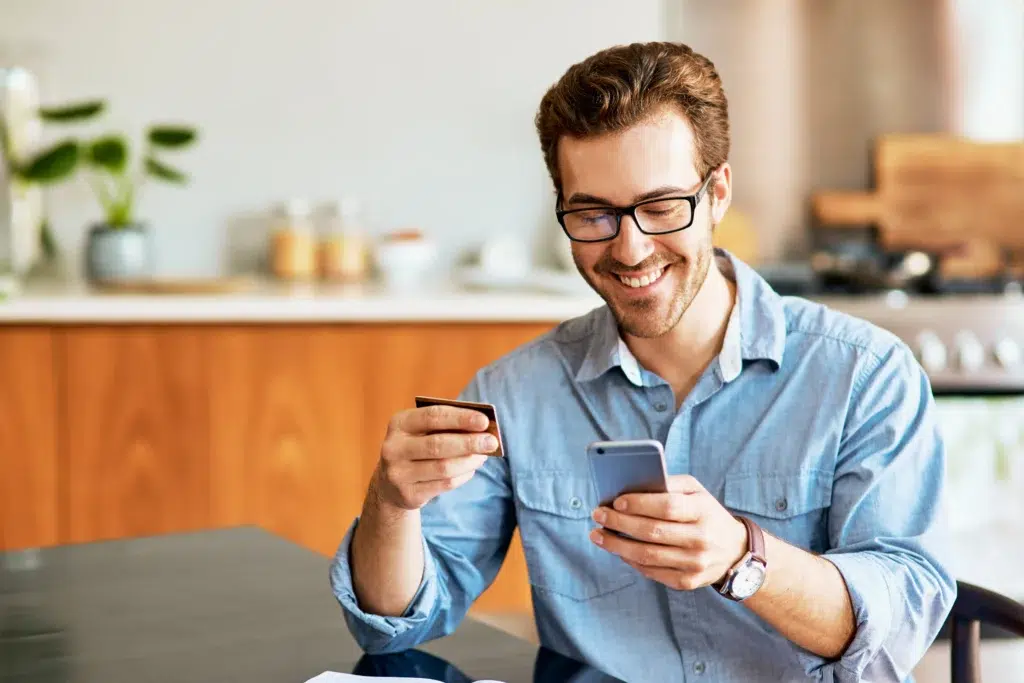 Homem de camisa azul, óculos de grau, sorrindo, segurando cartão e celular, ilustrando o que é sociedade de crédito direto.