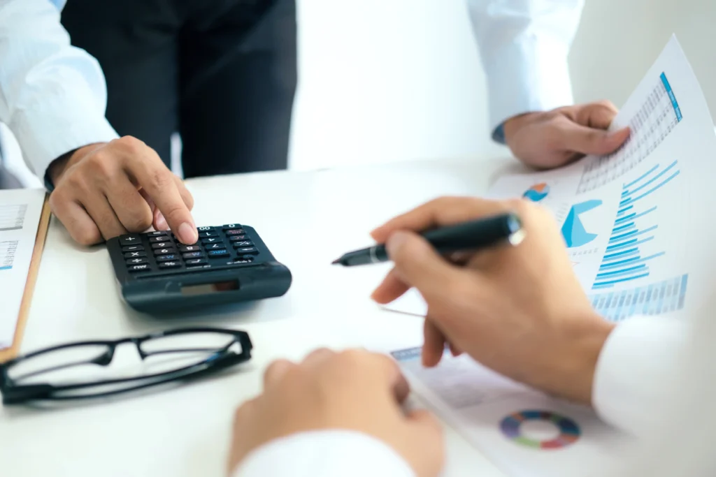 Mãos de duas pessoas fazendo organização financeira empresarial. Uma delas utiliza a calculadora e segura um papel com gráficos.