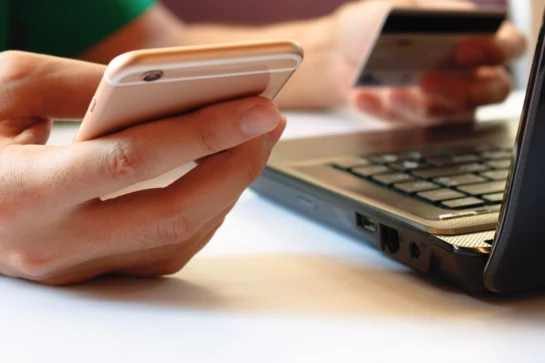 Pessoa usando celular e cartão de crédito ao lado de um laptop, representando como receber pagamento via link de forma rápida.