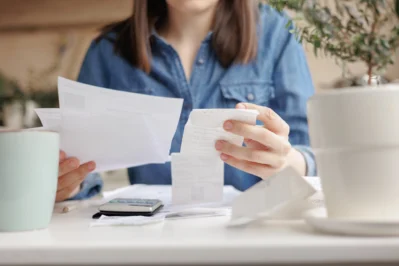 Mulher de camisa jeans, sentada em sofá, segurando papéis de contas e notas fiscais. Ela está calculando a possibilidade e vendo o que é amortização de dívida.