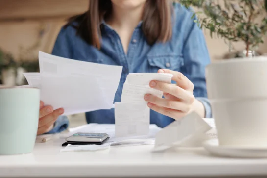 Mulher de camisa jeans, sentada em sofá, segurando papéis de contas e notas fiscais. Ela está calculando a possibilidade e vendo o que é amortização de dívida.