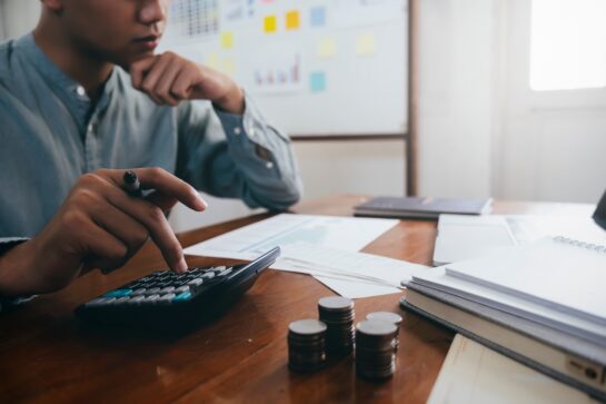 Homem usando calculadora ao lado de pilhas de moedas, com documentos ao fundo, simbolizando como receber por boleto.