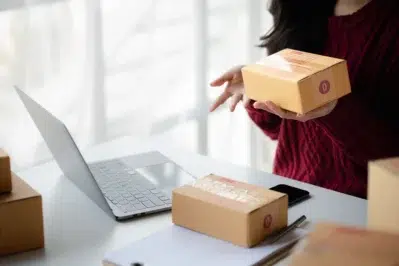 Mulher segurando caixas de entrega de pedidos online. Ela está aprendendo como vender produtos pela internet, assistindo vídeos no computador.