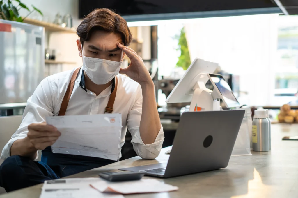 Homem sentado em balcão, segurando fatura e realizando o controle financeiro para restaurantes. Ele está com um computador em sua frente.