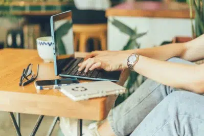 Mãos de uma pessoa usando um laptop em uma empresa, com itens de trabalho na mesa, utilizando internet banking empresarial.