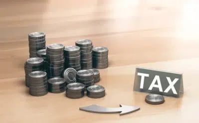 Imagem com pilhas de moedas metálicas ao lado de uma placa com a palavra "TAX", representando tributação, como IRPJ e CSLL.