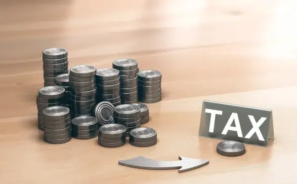 Imagem com pilhas de moedas metálicas ao lado de uma placa com a palavra "TAX", representando tributação, como IRPJ e CSLL.