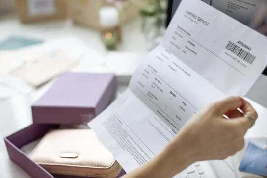 Mulher segura um papel enquanto tenta entender o que é nota fiscal de uma bolsa que está em cima de uma mesa. Ela lê sobre os dados de uma compra que realizou.