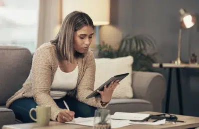 Empreendedora em sofá, com tablet na mão. Está anotando, em uma mesa de centro à sua frente, informações para os tipos de fluxo de caixa que fará para a empresa.