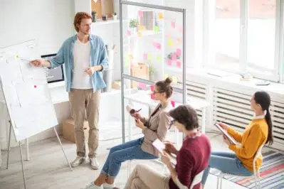 Trabalhadores em uma sala, enquanto homem está em pé apontando para dados em um papel. 3 mulheres estão sentadas em frente a ele, estudando como escalar um negócio.