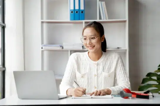 Mulher sorridente escreve em caderno diante de notebook, em escritório organizado. Ela está estudando como profissionalizar uma empresa.