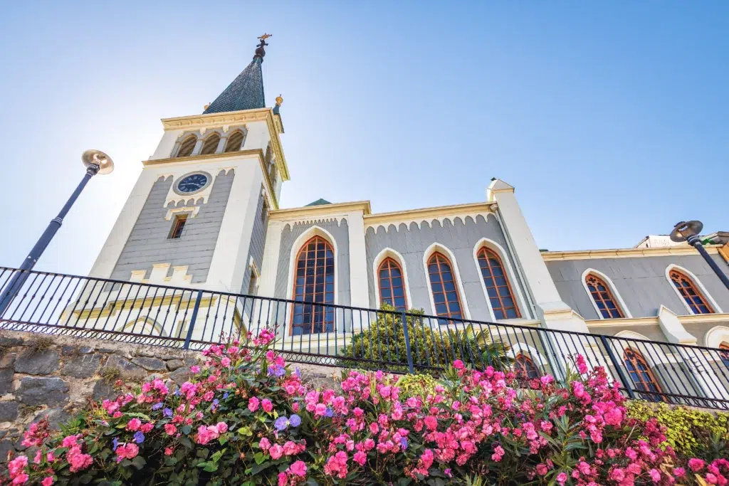 Igreja com torre e vitrais vista de baixo, flores no jardim em primeiro plano. Exemplo de inspiração para quem busca saber como abrir uma igreja.