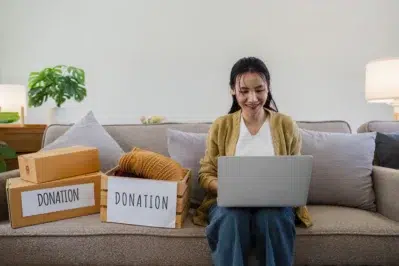 Mulher sentada no sofá com notebook, organizando doações em caixas e pesquisando sobre boleto proposta online.