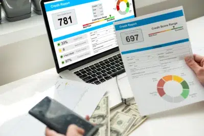Mãos segurando um papel e um celular. Na mesa, tem um notebook com uma análise de crédito, além de notas de dólar.