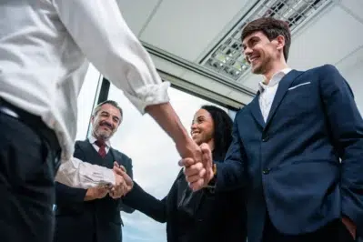 Empresários fechando acordo de aporte de capital após reuniões e contratos assinados. Todos os gestores estão reunidos em volta deles, que apertam as mãos.