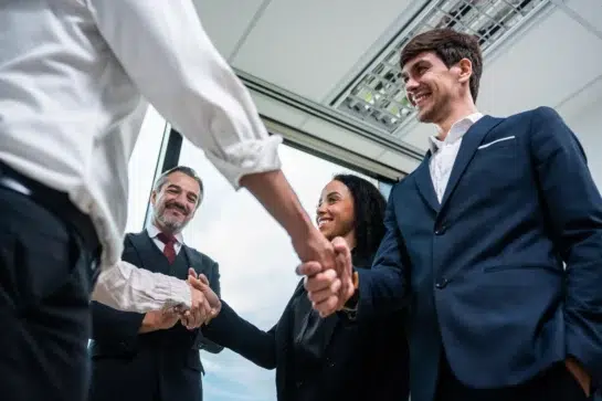 Empresários fechando acordo de aporte de capital após reuniões e contratos assinados. Todos os gestores estão reunidos em volta deles, que apertam as mãos.