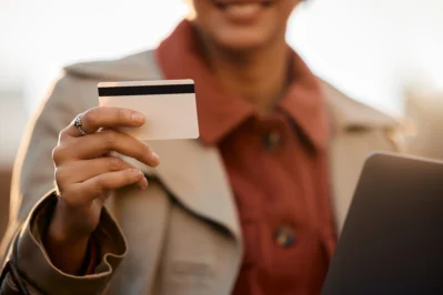 Mulher sorri enquanto segura um cartão de crédito pós-pago com a mão direita, em ambiente externo, usando notebook.