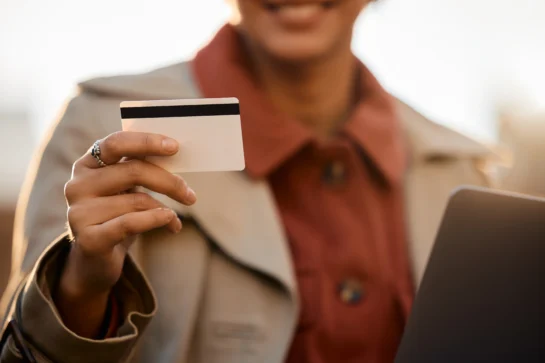 Mulher sorri enquanto segura um cartão de crédito pós-pago com a mão direita, em ambiente externo, usando notebook.