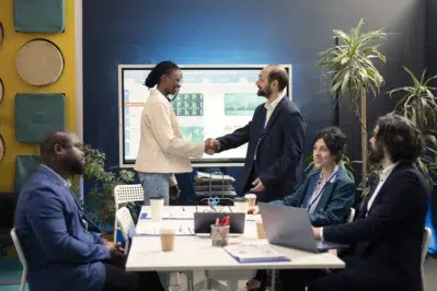 Pessoas em reunião apertando as mãos após acordo sobre compra e venda de empresas, com gráficos ao fundo em uma tela.