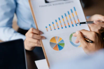 Pessoa analisa gráficos de indicadores financeiros em um relatório impresso, com barras e gráficos de pizza coloridos.
