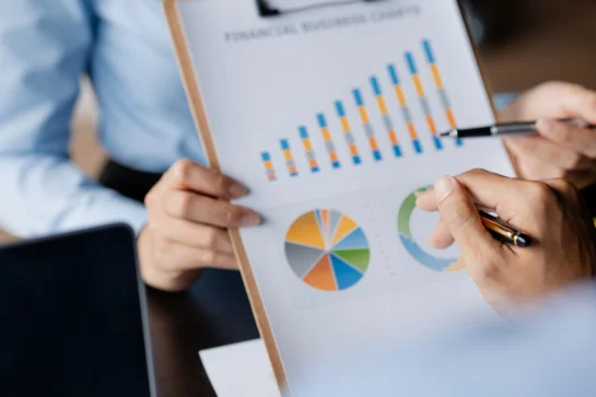 Pessoa analisa gráficos de indicadores financeiros em um relatório impresso, com barras e gráficos de pizza coloridos.