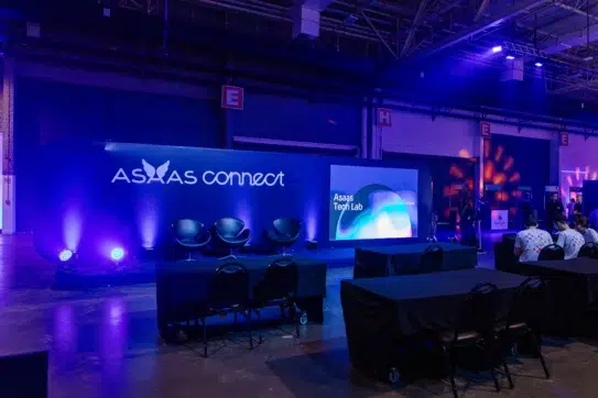 Espaço do Palco Connect com iluminação azul, três cadeiras no centro e tela lateral exibindo "Asaas Tech Lab", em ambiente de evento corporativo.