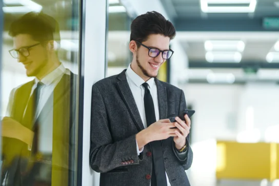 Empreendedor conferindo taxas Asaas pelo celular enquanto sorri em ambiente de escritório moderno.
