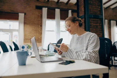 Como emitir boletos rápido sem conta bancária tradicional? Guia 9 Mulher em escritório, usando laptop e anotando em caderno procurando como emitir boletos rápido sem conta bancária tradicional.