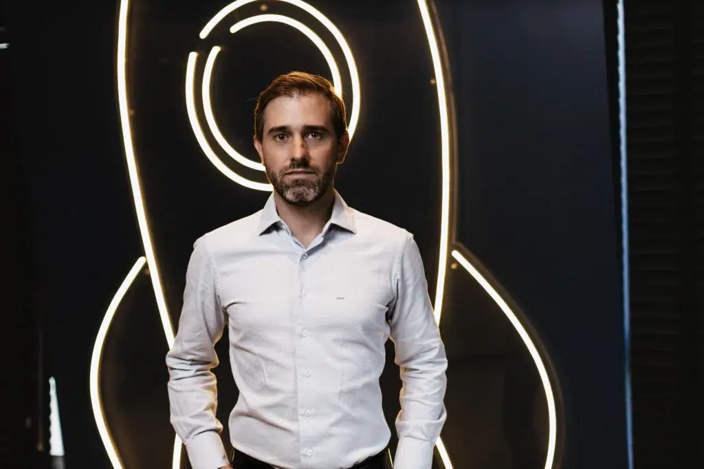 Foto de João Vitor Possamai, em frente a um fundo de led que desenha um foguete. Ele olha para foto e veste terno.
