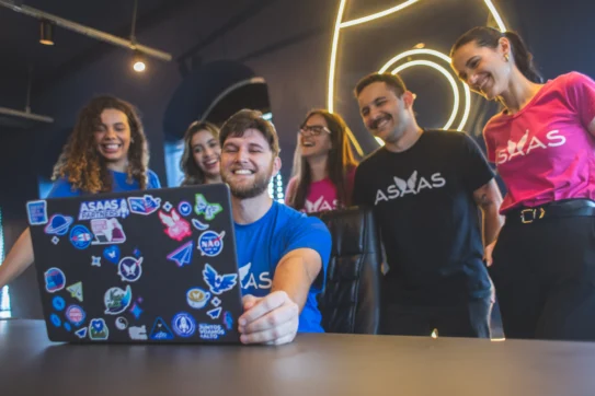 Equipe sorridente do Asaas reunida em frente a um notebook adesivado, representando o marco de 1000 tripulantes Asaas.