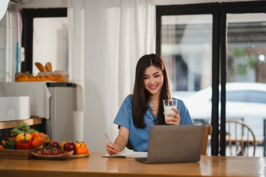 Mulher sorridente em frente ao notebook, tomando uma bebida e fazendo anotações sobre marketing para nutricionistas.