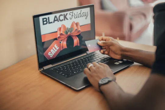 Pessoa segura um cartão de crédito em frente a um notebook com anúncio de promoções, destacando a segurança na Black Friday.
