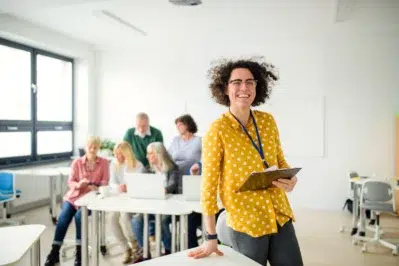 Professora sorridente com tablet em sala de aula, ilustrando como precificar o valor da mensalidade escolar.