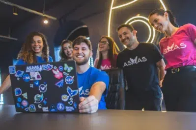 Imagem dos tripulantes reunidos em volta de um homem sentado numa cadeira, vendo a Retrospectiva Asaas 2025 no computador enquanto sorriem.