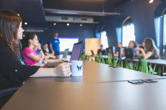 Colaboradora do Asaas segurando uma caneca em uma sala de reunião. Demais colaboradores conversam sobre as 3 aquisições do Asaas.