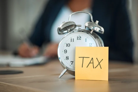 Relógio despertador com post-it escrito TAX representando obrigações fiscais do MEI e prazos tributários.