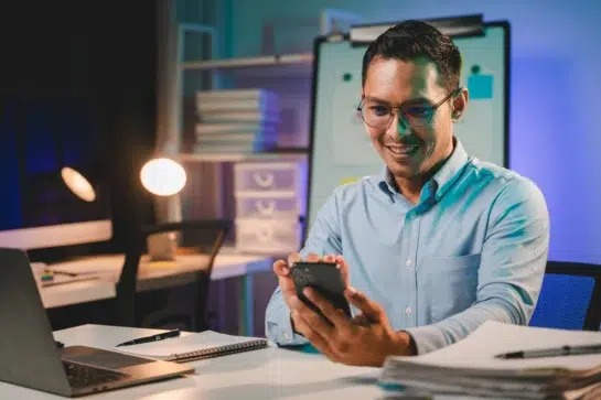 Homem sorrindo usando celular em escritório moderno, realizando pagamento com Pix via Open Finance de forma prática e segura.