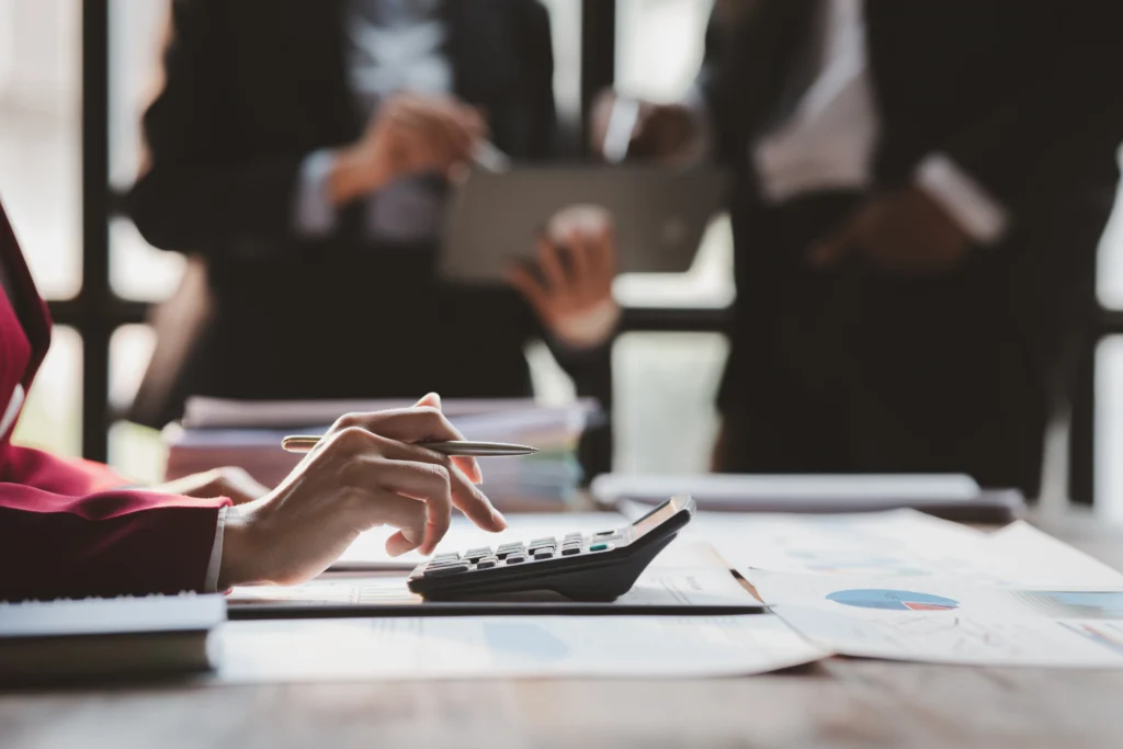 Como fazer cálculo de imposto sobre vendas online sem complicação? 4 Cálculo de imposto sobre vendas online sendo feito em calculadora com relatórios financeiros e equipe ao fundo.